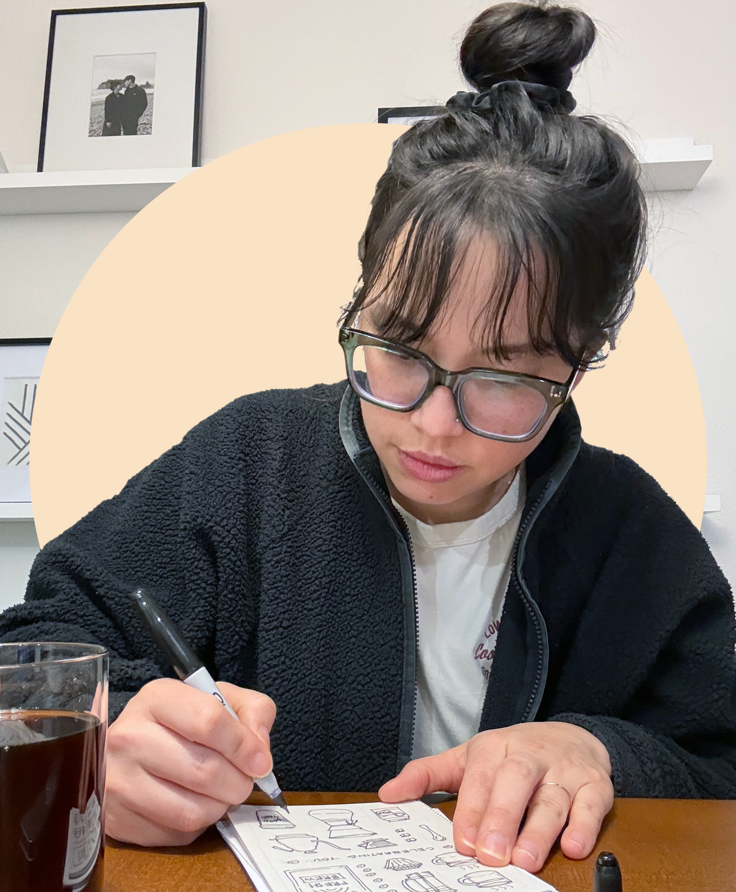 Person with glasses drawing on a piece of paper at a desk with a glass of coffee.